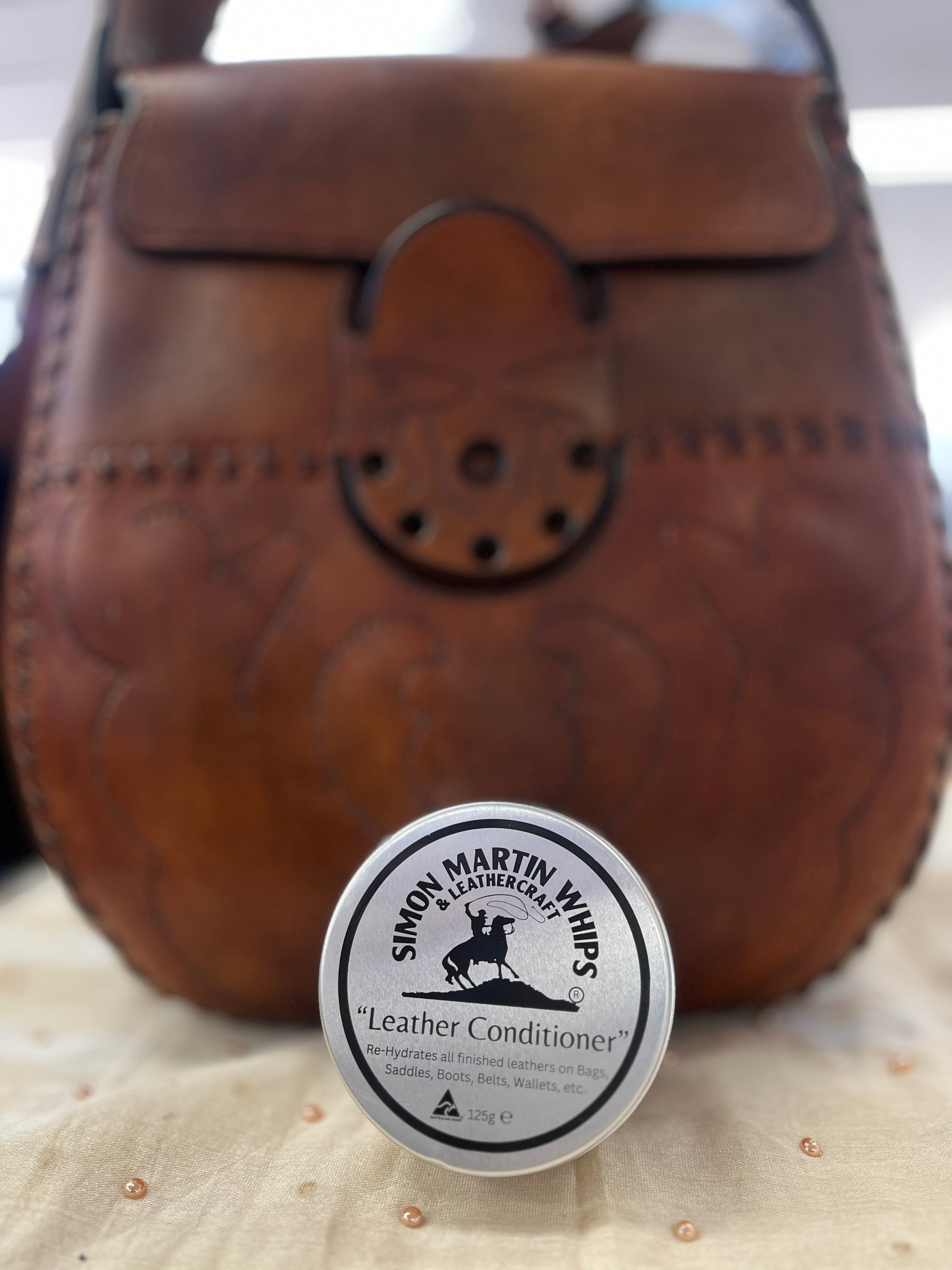Brown leather bag with a container of Simon Martin Whips leather conditioner on a beige surface.