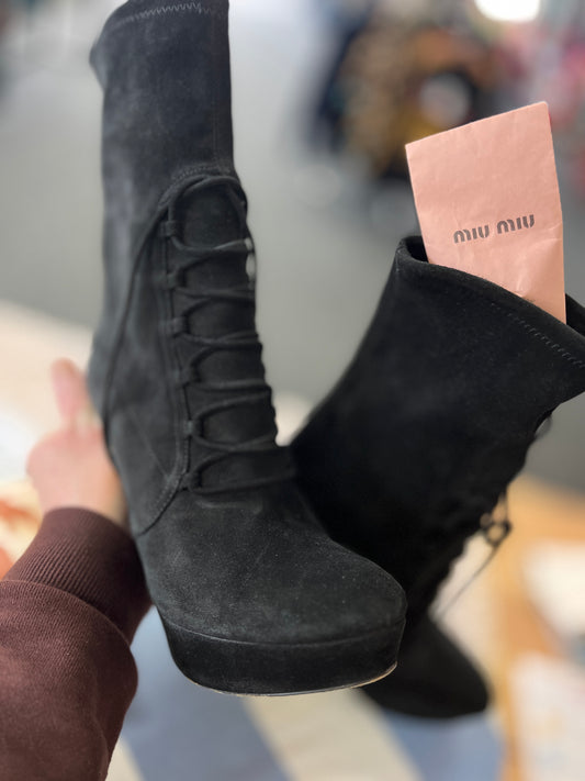 Black suede boots held by a person with a pink Miu Miu box in the background.